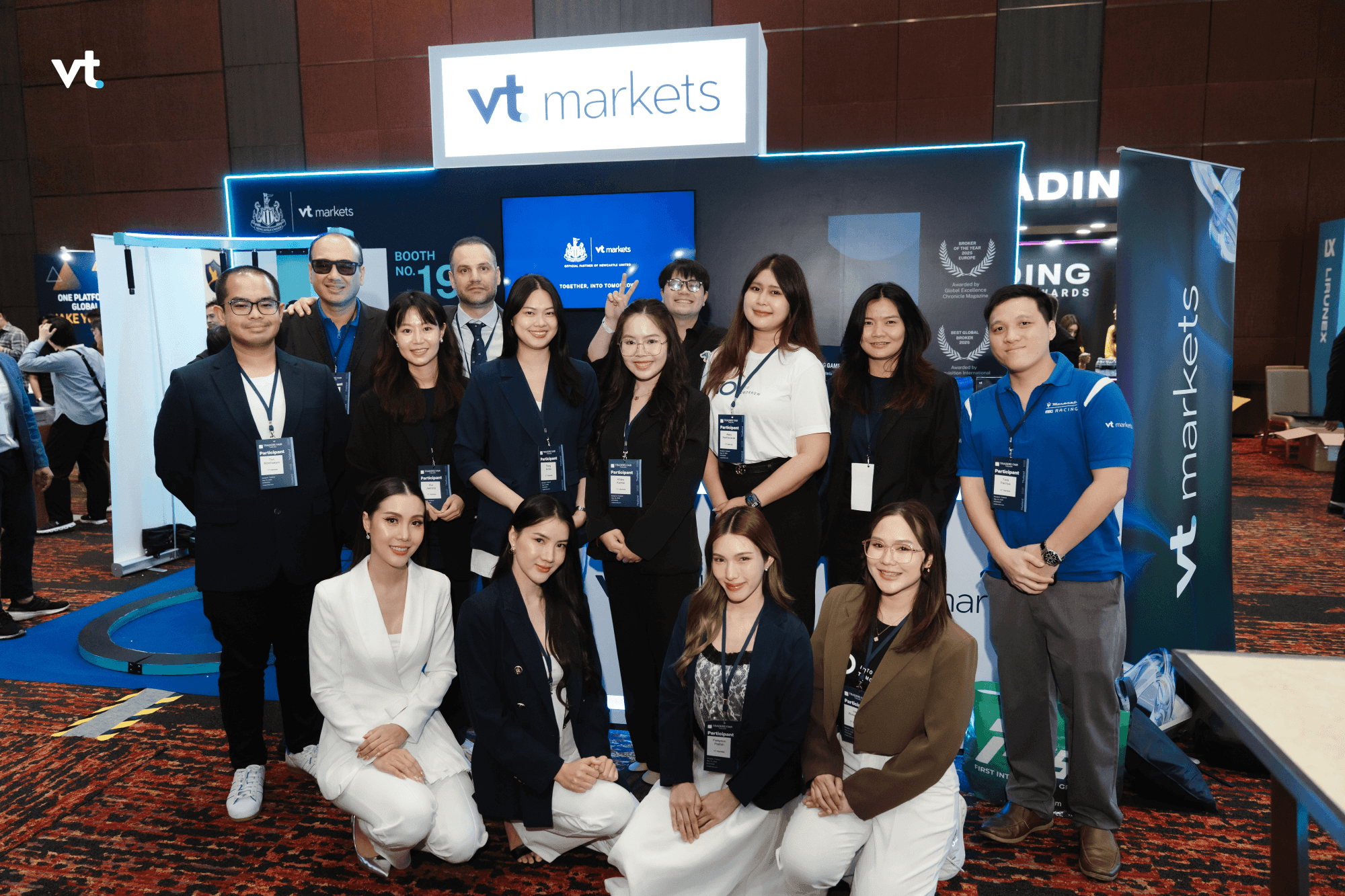 Group of professionals posing at a vt markets trade-show booth with blue branding and a large screen behind them